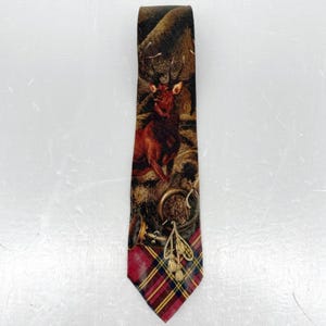 May include: A necktie featuring a detailed illustration of a brown stag in a forest setting, positioned above a gold-colored hunting horn with tassels. The bottom section of the tie displays a red, yellow, and black plaid tartan pattern.