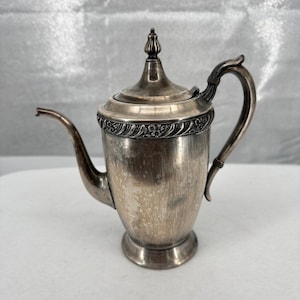 Op de afbeelding: Een zilverkleurige metalen koffiepot met een taps toelopend lichaam, een gebogen handvat en een lange, slanke tuit. Het deksel is bekroond met een knop en een decoratieve band met een bloemmotief in reliëf omcirkelt het bovenste deel.