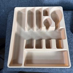 May include: A beige plastic cutlery tray with multiple compartments for organizing silverware. The tray has slots for knives, forks, spoons, and other utensils. The tray is rectangular and has a slightly textured surface.