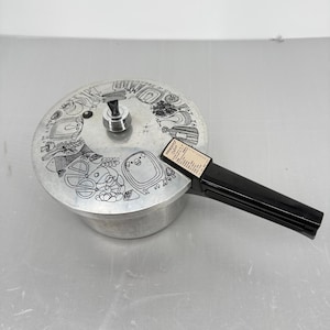 Puede incluir: Una olla a presión de aluminio plateado con mango negro y tapa circular con ilustraciones de línea negra de varios alimentos, incluyendo un cerdo, pescado y verduras. El mango muestra una etiqueta con el texto Presto Deluxe.