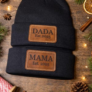 May include: Two black knit beanies with brown leather patches. One patch reads "DADA Est. 2025", the other reads "MAMA Est. 2025". The beanies are displayed on a wooden surface with holiday decorations, including pine cones and cinnamon sticks.