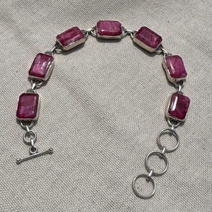 May include: A silver bracelet featuring rectangular, deep red gemstones. The gemstones are set in individual silver bezels and linked together with small silver chains. The bracelet has a toggle clasp closure.