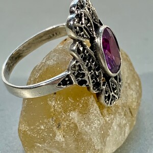 May include: A silver ring with an oval, deep purple gemstone. The ring has an ornate filigree design with small black accents. The band is thin and the ring is set on a light-colored stone.