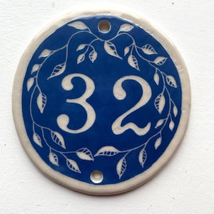 May include: A round, white ceramic house number sign with a blue background and white leafy vines. The number 32 is written in white on the blue background.