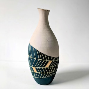 以下が含まれることがあります： クリーム色の部分と、金色の幾何学模様が施された濃い青色の部分がある、手作りのセラミック花瓶。 花瓶は細い首とわずかにフレアしたリムを持っています。 高さ約25cm。