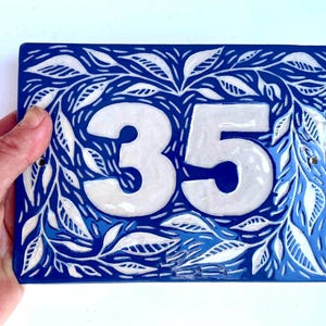 Könnte beinhalten: Rechteckige blaue Keramik-Hausnummerntafel mit der weißen Zahl 35. Die Tafel ist mit einem weißen Blattmuster um die Zahlen herum verziert. Die Tafel wird von einer Hand gehalten.