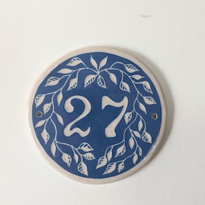 May include: Circular ceramic house number plaque featuring the number 27 in white on a blue base. The number is encircled by a white leaf pattern. The plaque has a cream border and two small holes for fixing.