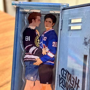 May include: A clear acrylic charm depicting two figures in hockey jerseys, one black with the number 81 and the other blue with the number 26. The charm is set inside a blue locker, with the text "HEATED RIVALS" visible.