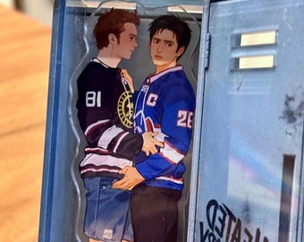 May include: A clear acrylic charm featuring two figures in hockey jerseys, one black with the number 81 and the other blue with the number 26. The charm is set inside a blue locker, with the text "HEATED RIVALS" visible.