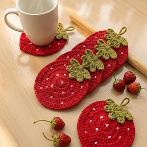 May include: A white mug sits on a red crocheted strawberry coaster. Several more strawberry coasters are arranged nearby, along with fresh strawberries. The coasters have green leaf accents and white seed details.