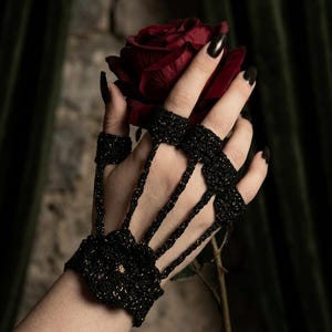 Può includere: Un accessorio per le mani all'uncinetto nero con dettagli dorati, composto da un polsino e anelli per le dita. L'accessorio viene mostrato mentre tiene una rosa rosso scuro. Lo sfondo include un muro strutturato e tessuto verde scuro.