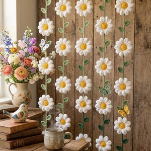 Può includere: Tenda di margherite all'uncinetto, con petali bianchi e centri gialli, infilata su steli verdi. La tenda è appesa davanti a un muro di legno. Le farfalle sono poste tra i fiori. Un vaso di fiori e un uccello adornano un tavolo di legno.