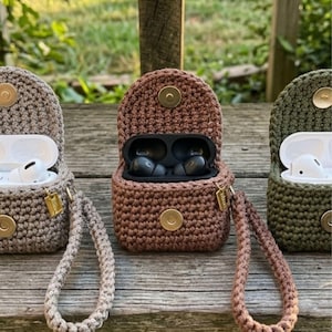 Peut inclure: Trois étuis pour écouteurs crochetés dans des tons de marron et de vert, chacun avec une dragonne assortie. Les étuis sont ouverts, révélant les écouteurs à l'intérieur. Les étuis ont un fermoir circulaire et une petite étiquette dorée.