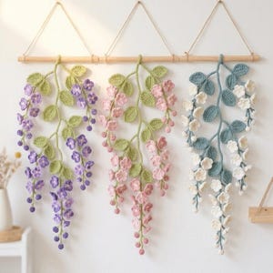 May include: Three crocheted wisteria wall hangings in purple, pink, and white. Each features cascading flowers and green leaves, suspended from a wooden dowel with twine. A charming, handmade floral decoration for the home.