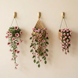 May include: Three crocheted hanging plant decorations suspended from gold-toned wall hooks. Each piece features green vines with small pink flowers. The center piece is in a tan woven basket, while the others are in green crocheted holders.