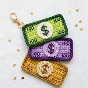 May include: Crocheted keychains in the shape of $100 bills in green, purple, and gold. Each bill features a crocheted dollar sign and the number 100. A gold-colored clasp is attached to the top bill.