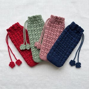 May include: Four handmade crochet drawstring pouches in red, green, pink, and navy blue. Each pouch has a textured stitch pattern and a matching drawstring with decorative flower-shaped ends. The pouches are displayed on a white linen surface.