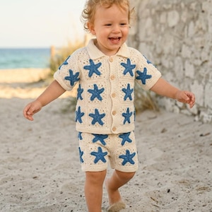 Può includere: Un completo all'uncinetto color crema composto da una camicia a maniche corte con bottoni e pantaloncini abbinati. Il capo presenta un motivo a quadrati ripetuti con un'applicazione a forma di stella marina blu scuro.