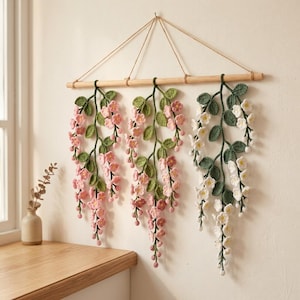 May include: A wall hanging featuring three crocheted floral vines suspended from a horizontal wooden dowel. Two vines display pink blossoms with green leaves, while the third vine features white flowers with green leaves, all crafted from yarn.