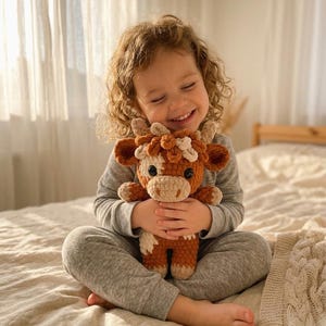 May include: A crocheted brown and white cow plush toy with large eyes and small horns. The plush is being held by a person wearing gray pajamas. The cow has a light brown muzzle and a white patch on its forehead. The toy is handmade.