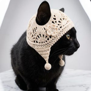 Puede incluir: Un gato negro con un sombrero de ganchillo beige con detalles florales y calados. El sombrero tiene dos solapas colgantes para las orejas con pequeños pompones. El gato está sentado sobre una superficie de mármol.