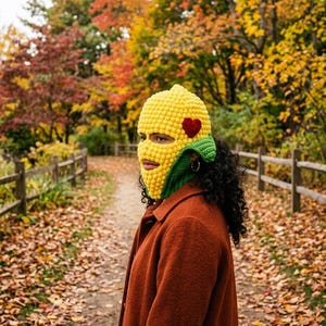 May include: A yellow crochet balaclava with a red heart accent and green trim. The balaclava is worn with a rust-colored coat. The background features a path with fallen leaves and trees with autumn foliage.