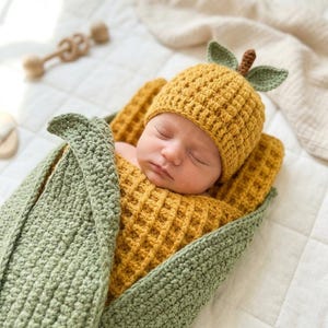 Può includere: Un neonato avvolto in una coperta all'uncinetto e con un cappello abbinato. Il cappello è giallo con dettagli di foglie verdi e la coperta è verde e gialla. Il bambino dorme pacificamente su una superficie bianca.