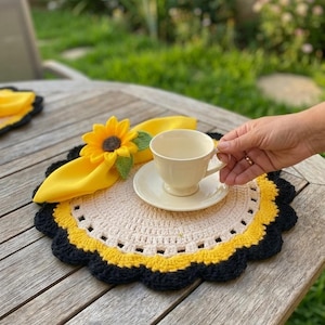 May include: A round, crocheted placemat featuring a cream-coloured centre, a yellow middle ring, and a scalloped black border. A bright yellow fabric napkin adorned with a three-dimensional sunflower and green leaf sits atop the placemat.
