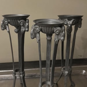 Puede incluir: Tres soportes para plantas antiguos de color plateado. Cada soporte tiene una parte superior redonda sostenida por tres patas. Las patas están adornadas con detalles de cabeza de carnero. Los soportes se exhiben en una bandeja plateada.