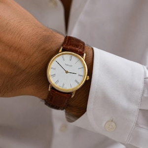 May include: A Tissot wristwatch featuring a round gold-tone case, a white dial with black Roman numeral hour markers, and a brown leather strap with a textured crocodile-embossed pattern. The watch has thin black hands and a simple, clean design.
