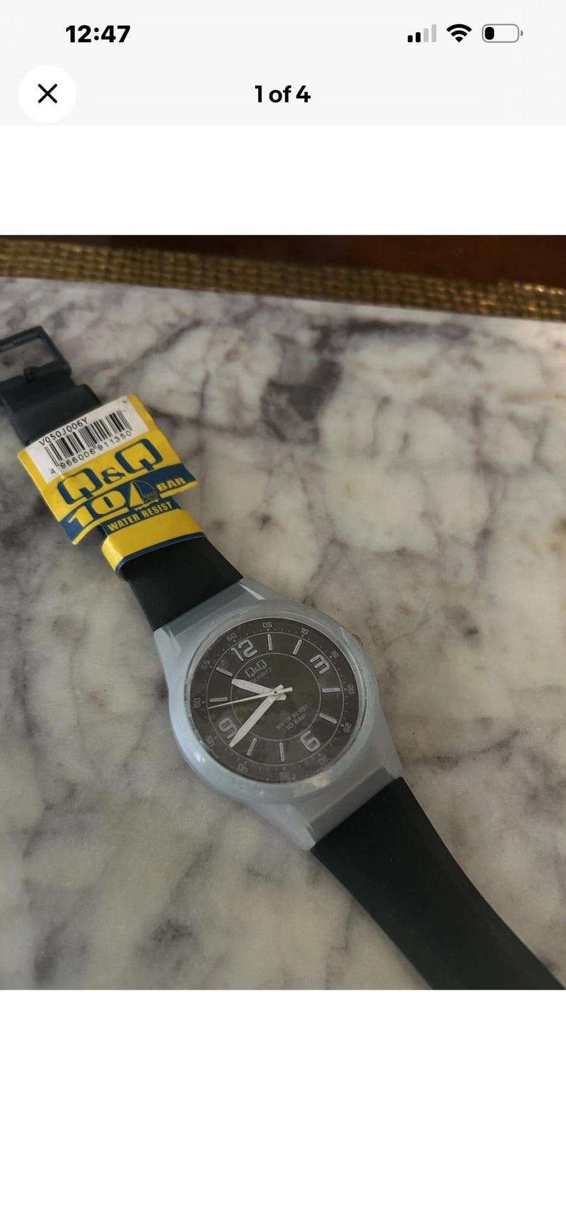 Puede incluir: Un reloj de pulsera de cuarzo Q&Q con correa de silicona negra y caja gris claro. La esfera negra presenta n&uacute;meros ar&aacute;bigos blancos en las posiciones 12, 3, 6 y 9, con manecillas blancas. Una etiqueta amarilla y azul indica 10 BAR.