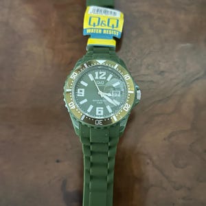 Puede incluir: Un reloj de pulsera de cuarzo Q&Q en color verde oliva con correa de silicona a juego y bisel giratorio. La esfera muestra n&uacute;meros ar&aacute;bigos blancos en las 6, 9 y 12, una ventana de fecha en la posici&oacute;n de las 3 y el texto Q&Q, QUARTZ, WATER RESIST 5 BAR.