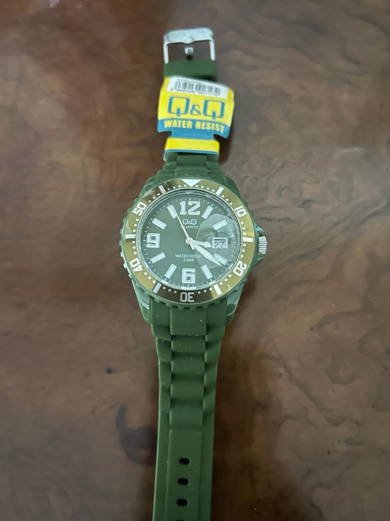 Puede incluir: Un reloj de pulsera de cuarzo Q&Q en color verde oliva con correa de silicona a juego y bisel giratorio. La esfera muestra n&uacute;meros ar&aacute;bigos blancos en las 6, 9 y 12, una ventana de fecha en la posici&oacute;n de las 3 y el texto Q&Q, QUARTZ, WATER RESIST 5 BAR.