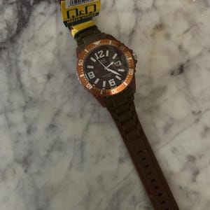 Puede incluir: Un reloj de pulsera Q&Q marr&oacute;n con esfera marr&oacute;n oscuro, marcadores de hora blancos y un bisel giratorio con detalles en naranja. El reloj incluye una correa de silicona marr&oacute;n y una etiqueta amarilla sujeta a la banda.