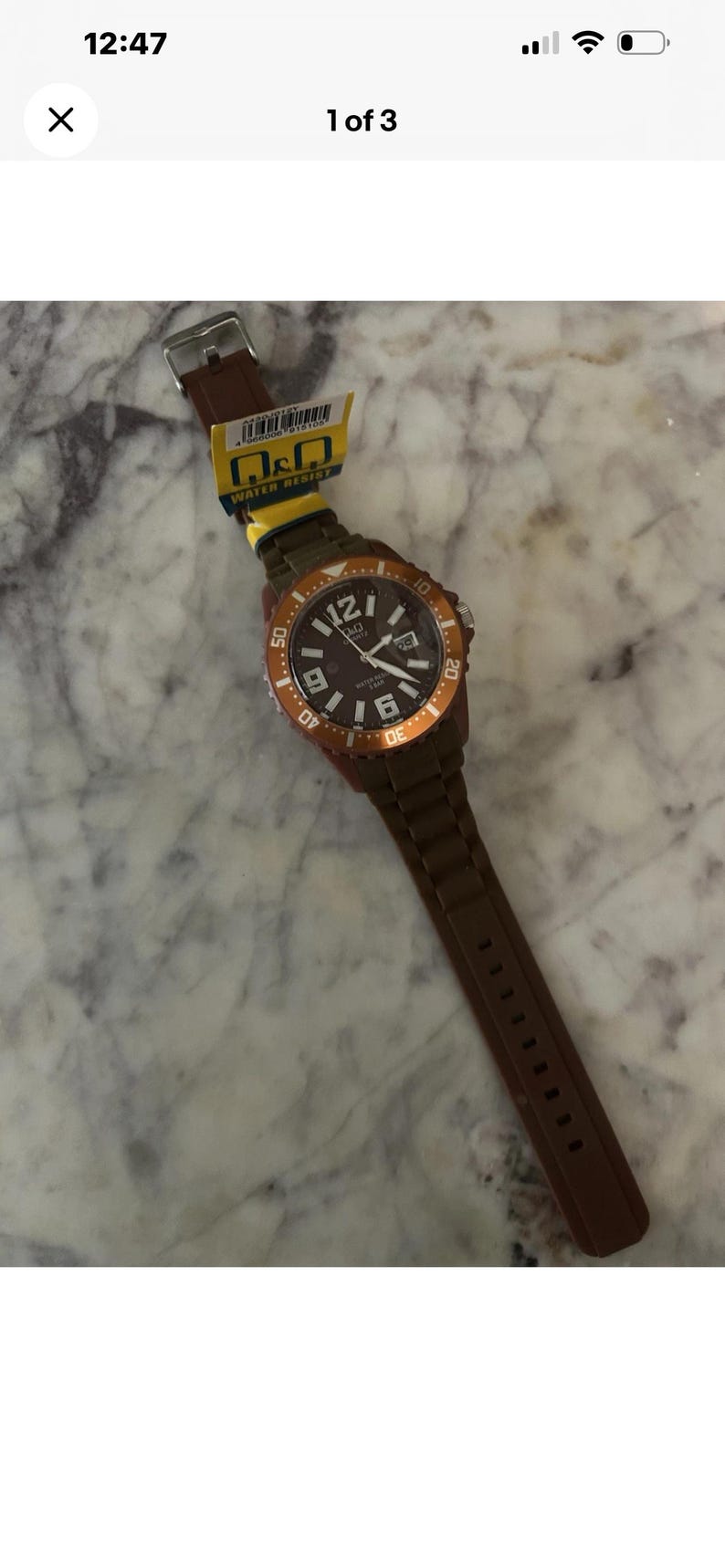 Puede incluir: Un reloj de pulsera Q&Q marr&oacute;n con esfera marr&oacute;n oscuro, marcadores de hora blancos y un bisel giratorio con detalles en naranja. El reloj incluye una correa de silicona marr&oacute;n y una etiqueta amarilla sujeta a la banda.