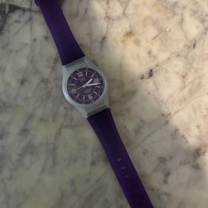 Puede incluir: Un reloj de pulsera de cuarzo Q&Q con una correa de silicona de color morado vibrante y una esfera circular morada a juego. El reloj tiene una caja de pl&aacute;stico gris claro, marcadores de hora blancos y manecillas blancas. La esfera muestra el 12 y el 6.