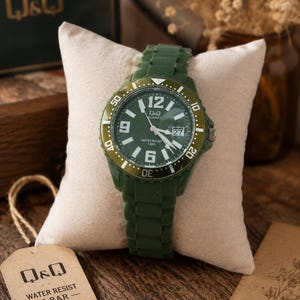Puede incluir: Un reloj de pulsera de cuarzo Q&Q con correa de silicona verde oliva oscuro y esfera verde a juego. El reloj presenta n&uacute;meros ar&aacute;bigos blancos en las 6, 9 y 12, una ventana de fecha a las 3 y un bisel giratorio. Una etiqueta dice Q&Q WATER RESIST 5 BAR.