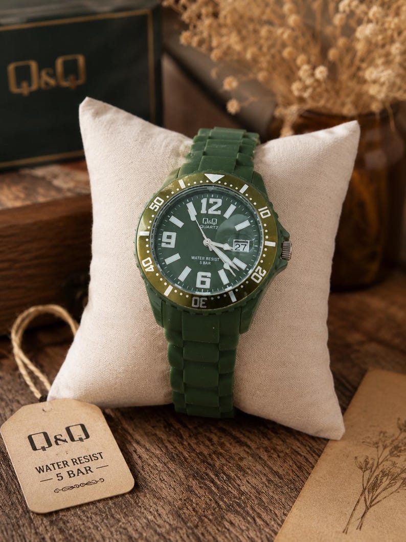 Puede incluir: Un reloj de pulsera de cuarzo Q&Q con correa de silicona verde oliva oscuro y esfera verde a juego. El reloj presenta n&uacute;meros ar&aacute;bigos blancos en las 6, 9 y 12, una ventana de fecha a las 3 y un bisel giratorio. Una etiqueta dice Q&Q WATER RESIST 5 BAR.
