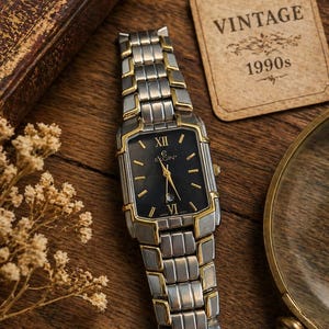 Puede incluir: Reloj Elgin vintage de los años 90. El reloj tiene una esfera negra rectangular con detalles dorados y una correa plateada y dorada. El reloj se exhibe sobre una superficie de madera con una etiqueta vintage.