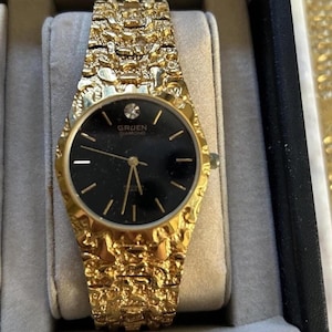 May include: A gold-coloured Gruen Diamond watch with a textured strap and a black watch face. The watch face has gold hands and markers, and the brand name is visible. The watch is presented in a black box with a grey interior.