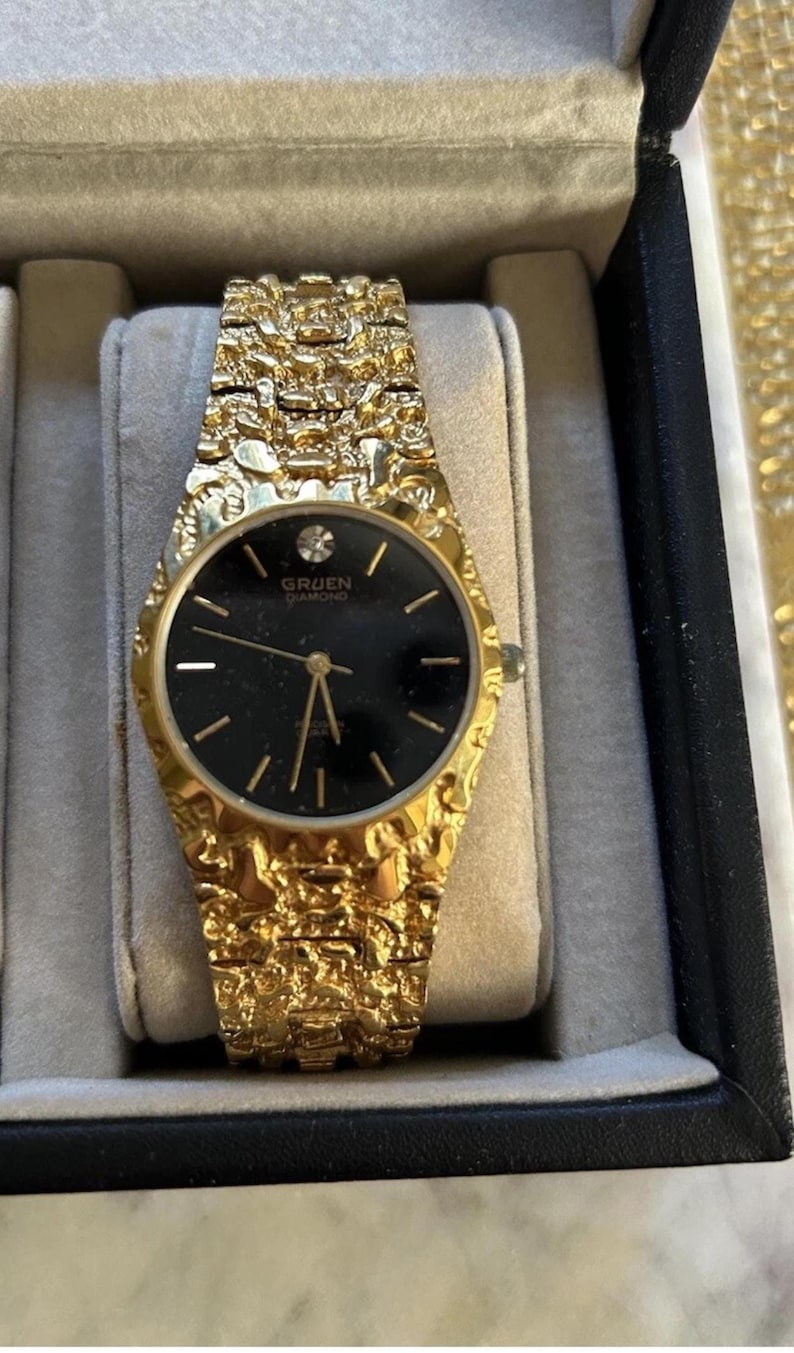 May include: A gold-coloured Gruen Diamond watch with a textured strap and a black watch face. The watch face has gold hands and markers, and the brand name is visible. The watch is presented in a black box with a grey interior.