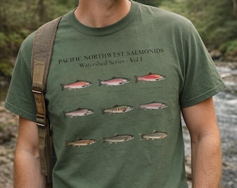 May include: Green t-shirt featuring illustrations of various salmon species, including Chinook, Coho, and Sockeye. The shirt has the text "PACIFIC NORTHWEST SALMONIDS Watershed Series - Vol 1" printed above the fish illustrations. The shirt is being worn by a person.