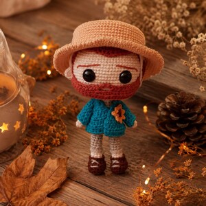 Puede incluir: Una muñeca de ganchillo con sombrero color canela, barba roja y ojos de botón negros. La muñeca viste una chaqueta azul con un pequeño detalle de girasol naranja en la solapa, pantalones claros y zapatos marrón oscuro, con puntos visibles.