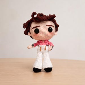 Puede incluir: Una muñeca de ganchillo que representa una figura con cabello rizado de color marrón oscuro, ojos negros grandes y mejillas rosadas. La muñeca viste una camisa rosa de manga larga con cuello y pantalones blancos acampanados con detalles dorados.