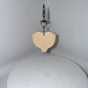 Op de afbeelding: Een beige leren sleutelhanger met een decoratieve vorm, opgehangen aan een zilverkleurige metalen clip. De sleutelhanger heeft een uniek, gestileerd ontwerp. De achtergrond is lichtgrijs.