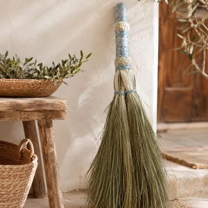 Può includere: Una scopa a manico lungo con un fascio di setole verdi naturali e rigide alla base. Il manico è avvolto da una combinazione di corda intrecciata azzurra e bianca, intervallata da sezioni di fibra naturale intrecciata di colore beige.