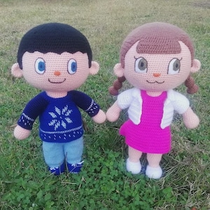 May include: Two handmade crochet dolls, a boy and a girl, stand side-by-side on green grass. The boy doll wears a blue sweater with a snowflake design and blue pants. The girl doll wears a pink dress and a white jacket. Both dolls have large eyes and are holding hands.