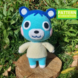 Peut inclure: Poupée ours bleue au crochet avec un museau, des oreilles et des pattes blanches, et un pull crème. La poupée a de grands yeux noirs et des joues roses. Le texte "PATTERN BY CROCHETCONI" est dans un encadré vert. La poupée est posée sur une souche de bois.