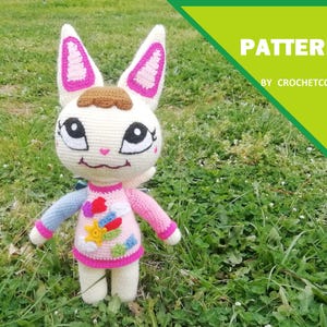 Puede incluir: Muñeca de ganchillo hecha a mano con cuerpo blanco, orejas rosas y una mancha marrón en la cabeza. La muñeca lleva una camisa rosa con parches coloridos y mangas azules y rosas. La palabra "PATTERN" es visible en un cuadro verde.