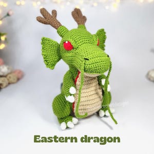 May include: A green crocheted dragon plush toy featuring brown antlers, bright red eyes, and a beige belly. The creature has long, thin whiskers extending from its snout, white claws on its feet, and textured green wings. The text Eastern dragon is below.